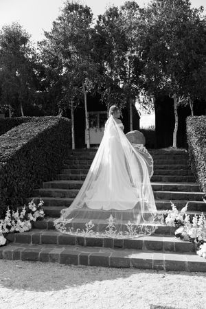 Black and white photo of a bride ascending stone steps in a flowing gown with an embroidered cathedral veil.