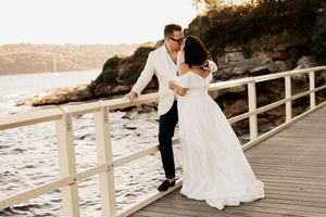 Bride and groom share a romantic kiss on a waterfront pier, dressed in elegant white wedding attire with ocean views.