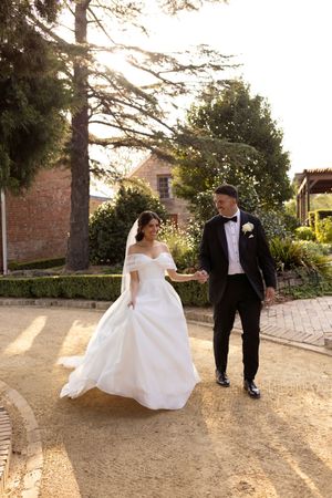 Newlyweds strolling through golden sunlit garden, bride in elegant white ballgown, groom in classic black tuxedo.