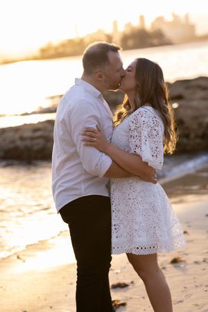 Couple sharing a romantic kiss on the beach at golden sunset, woman in white lace dress, city skyline in background.