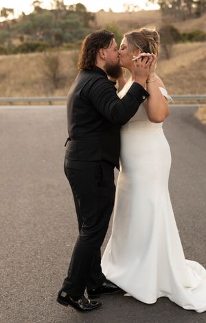 Bride and groom sharing a romantic kiss on a road, wearing white gown and black suit, with scenic hills at sunset.