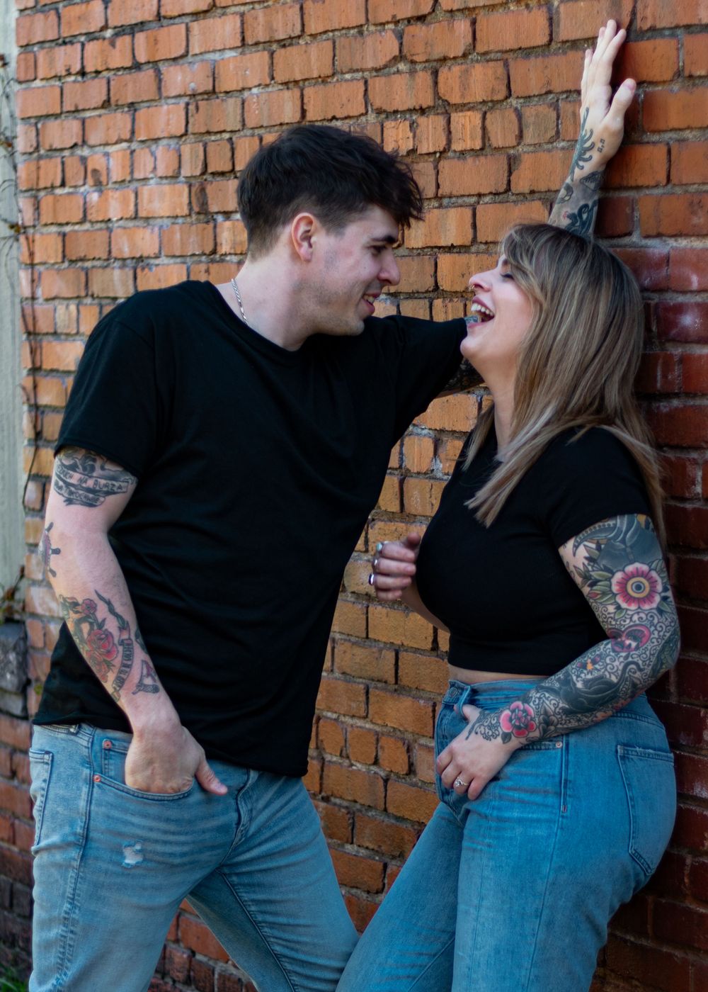 Couple in black shirts and jeans posing against rustic brick wall.