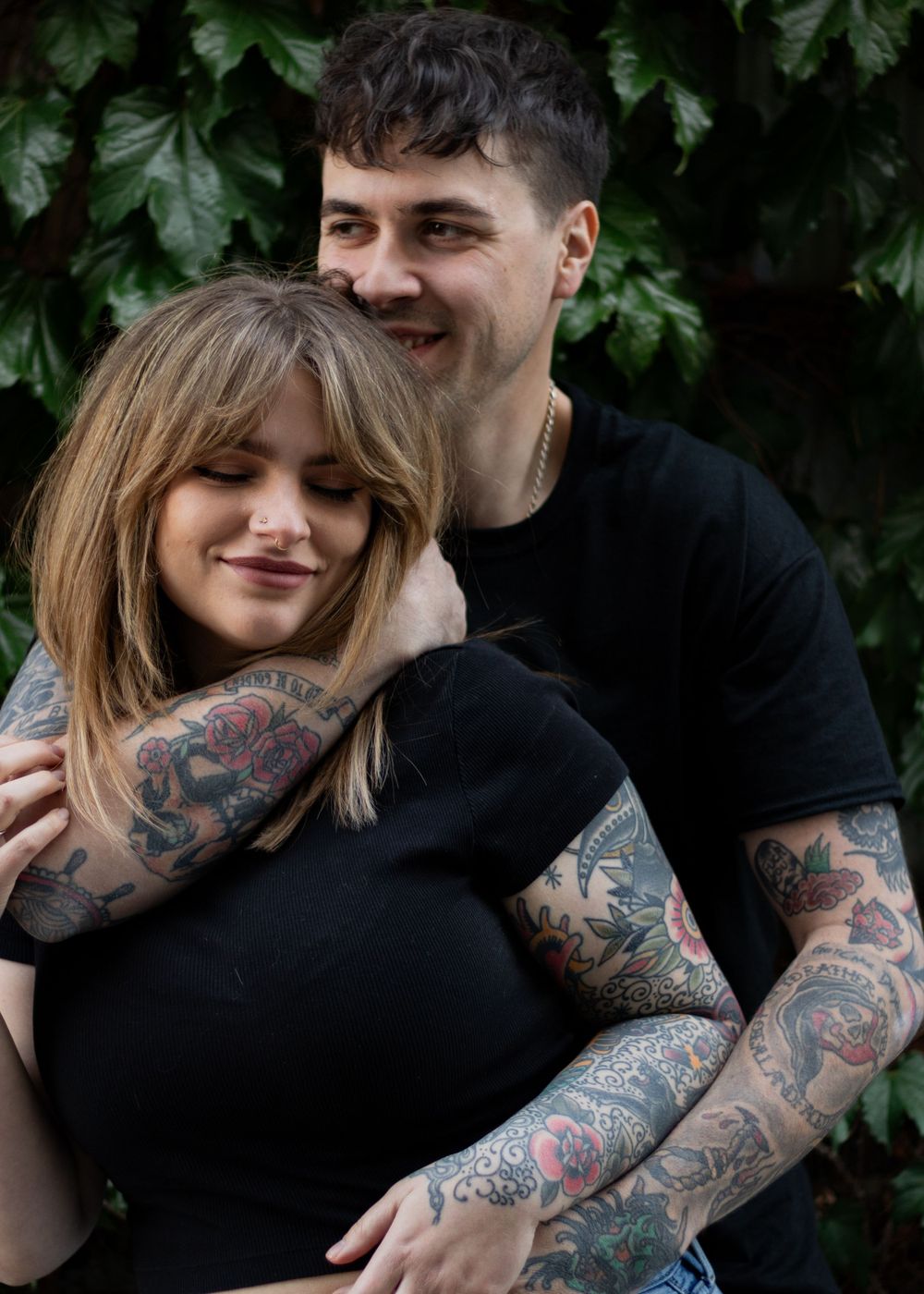Couple embracing with colorful tattoo sleeve visible against green foliage.