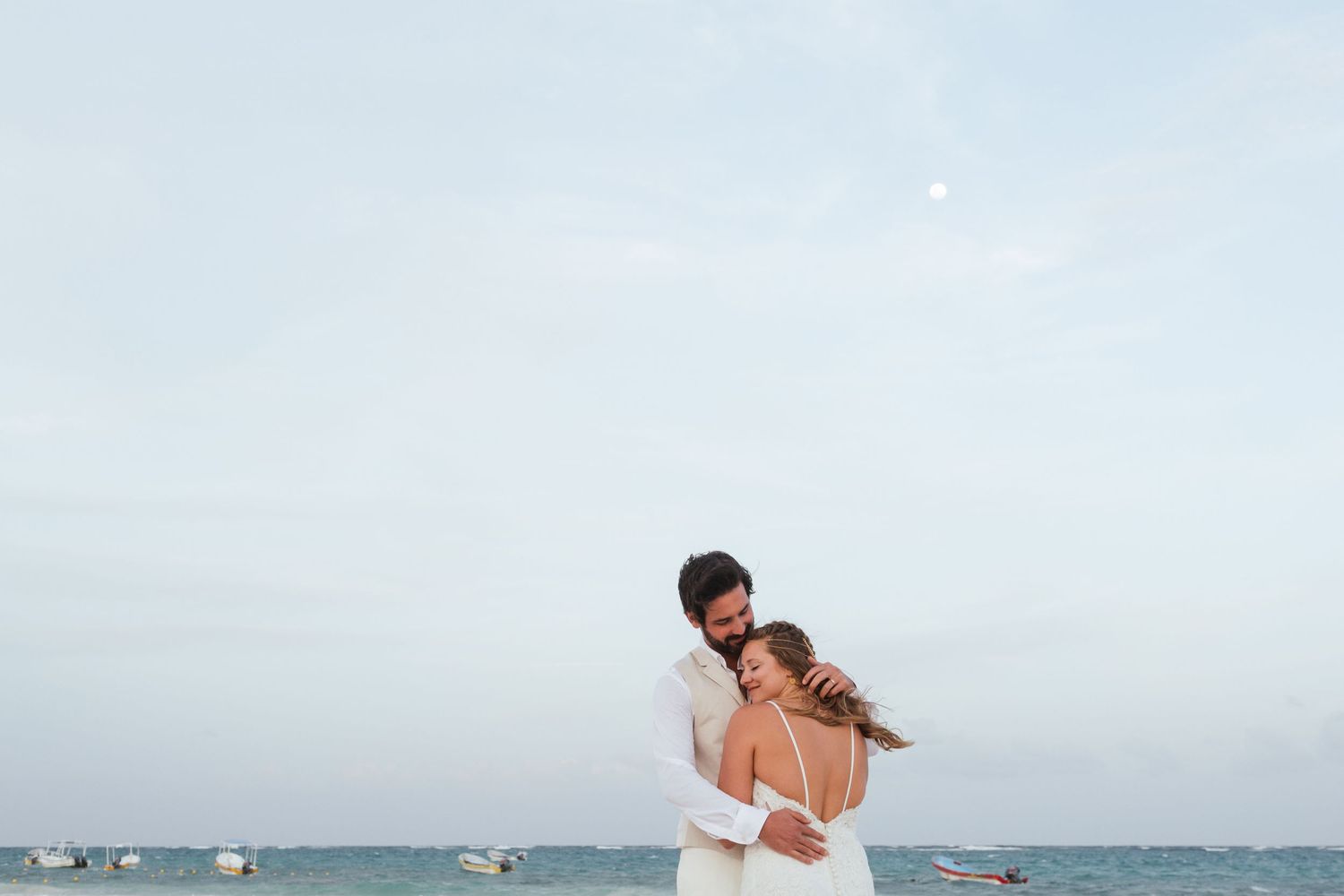 A couple in wedding attire embraces on a beach with calm ocean waters and boats in the background.