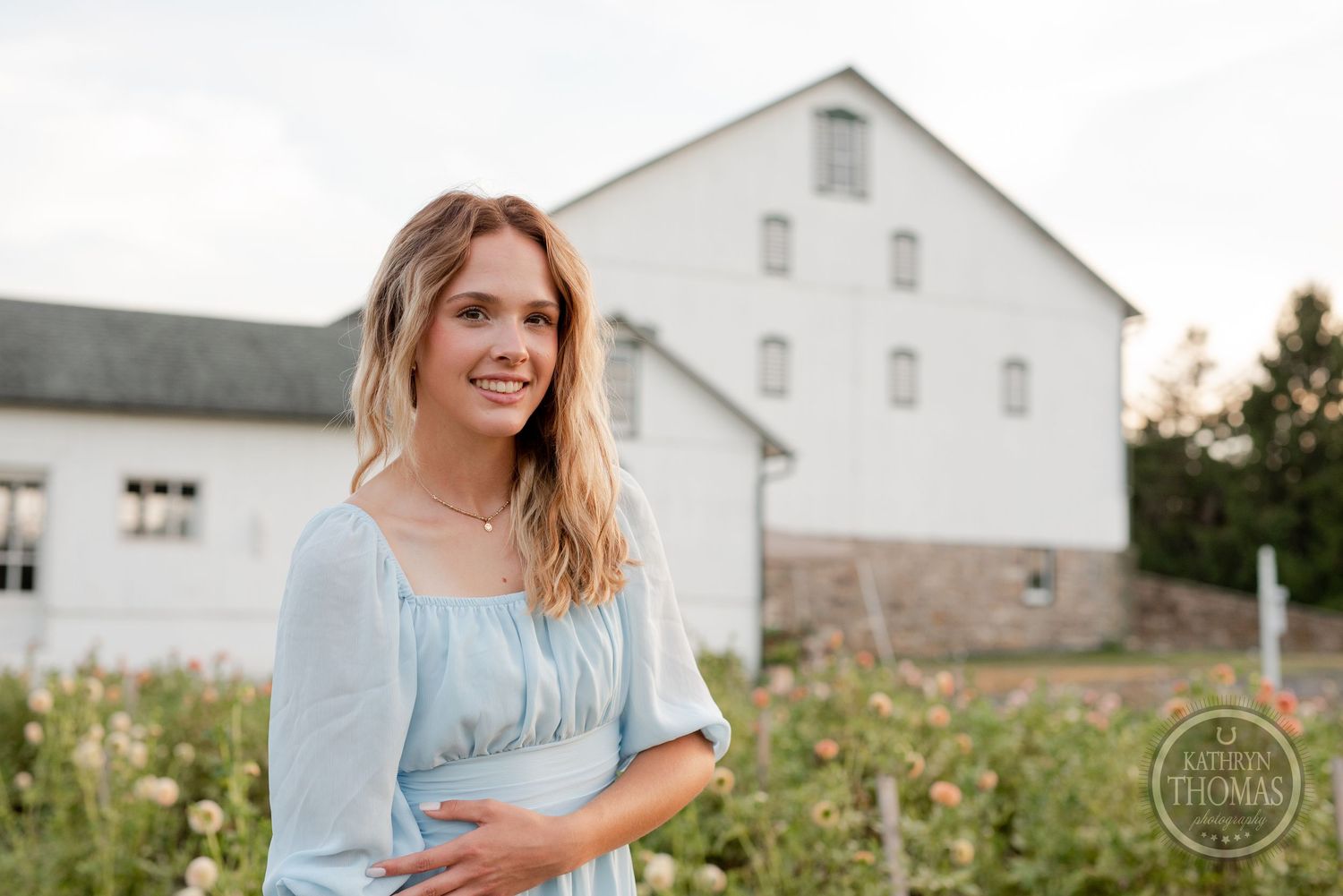 A summery portrait features a light blue dress against a rustic white barn and flowering garden setting.