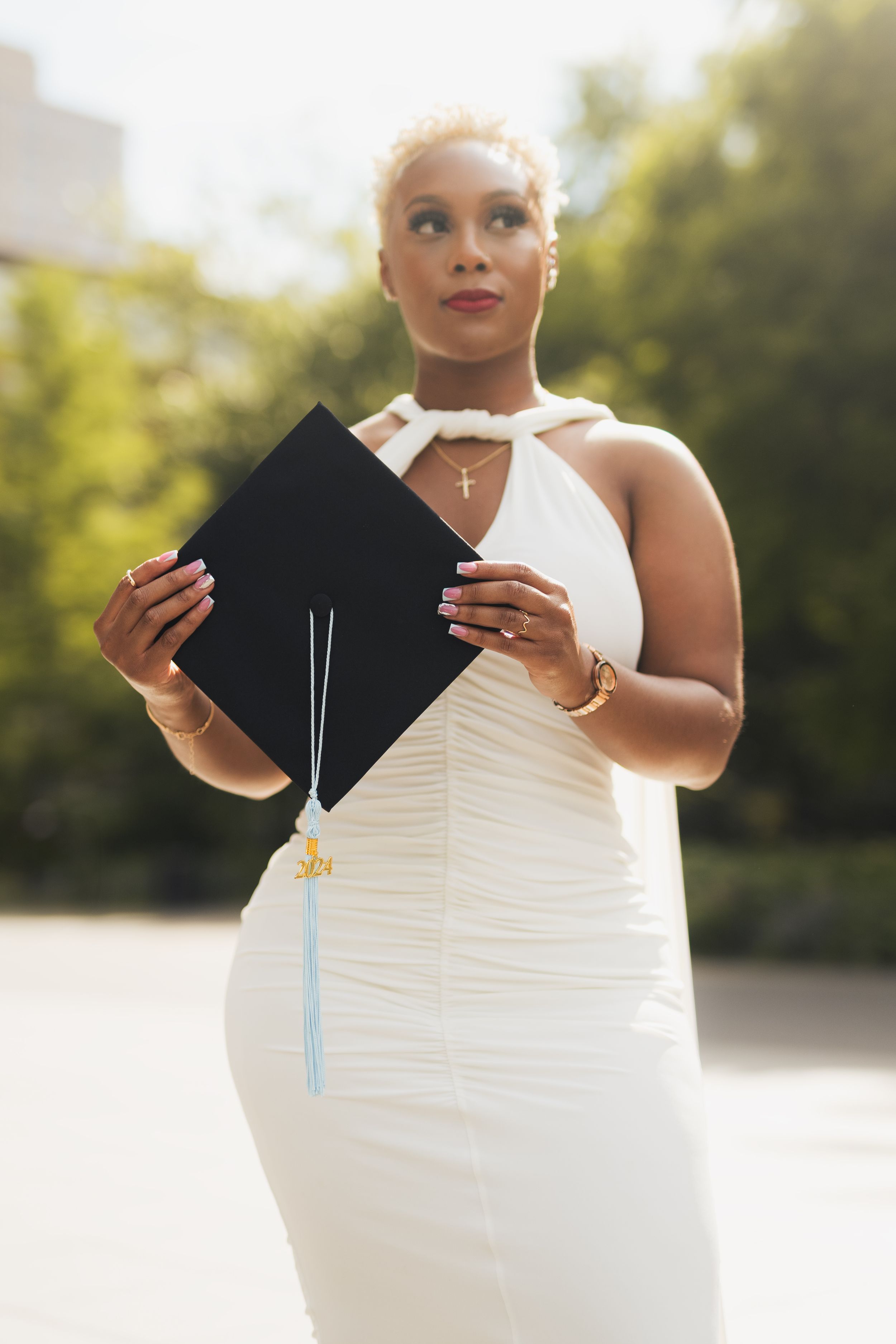 Person in white formal dress holding graduation cap against bright outdoor backdrop.