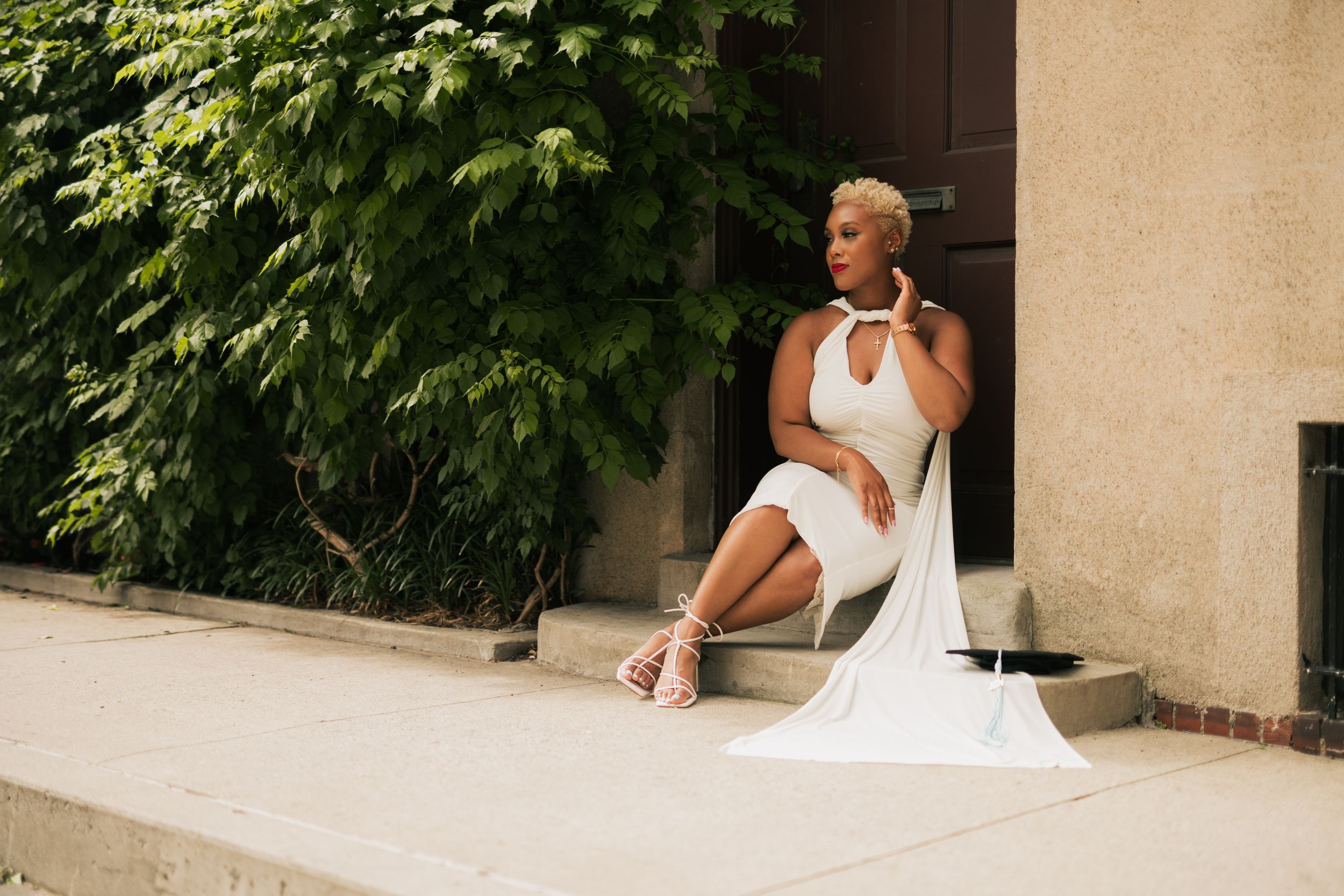 Person in white dress seated outdoors against natural greenery and beige wall background.