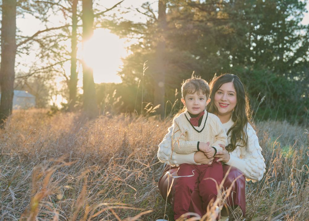 Two people sit together in tall grass during golden hour with sunlight filtering through trees.