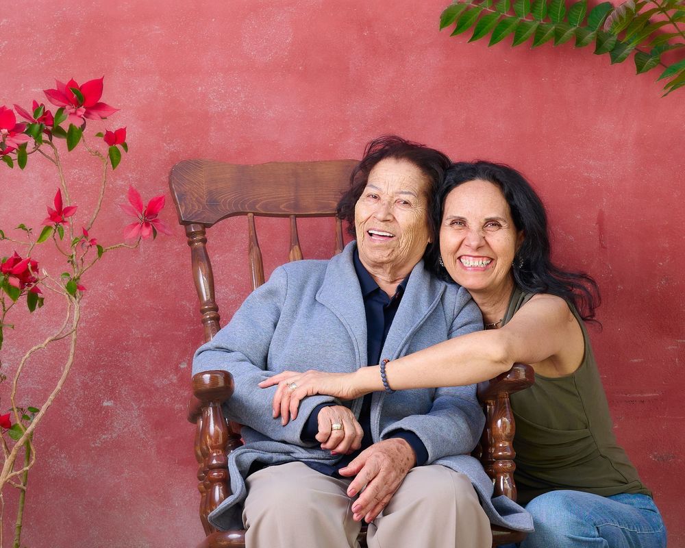 Two people smile while sitting together on a wooden chair against a pink wall with flowering vines.