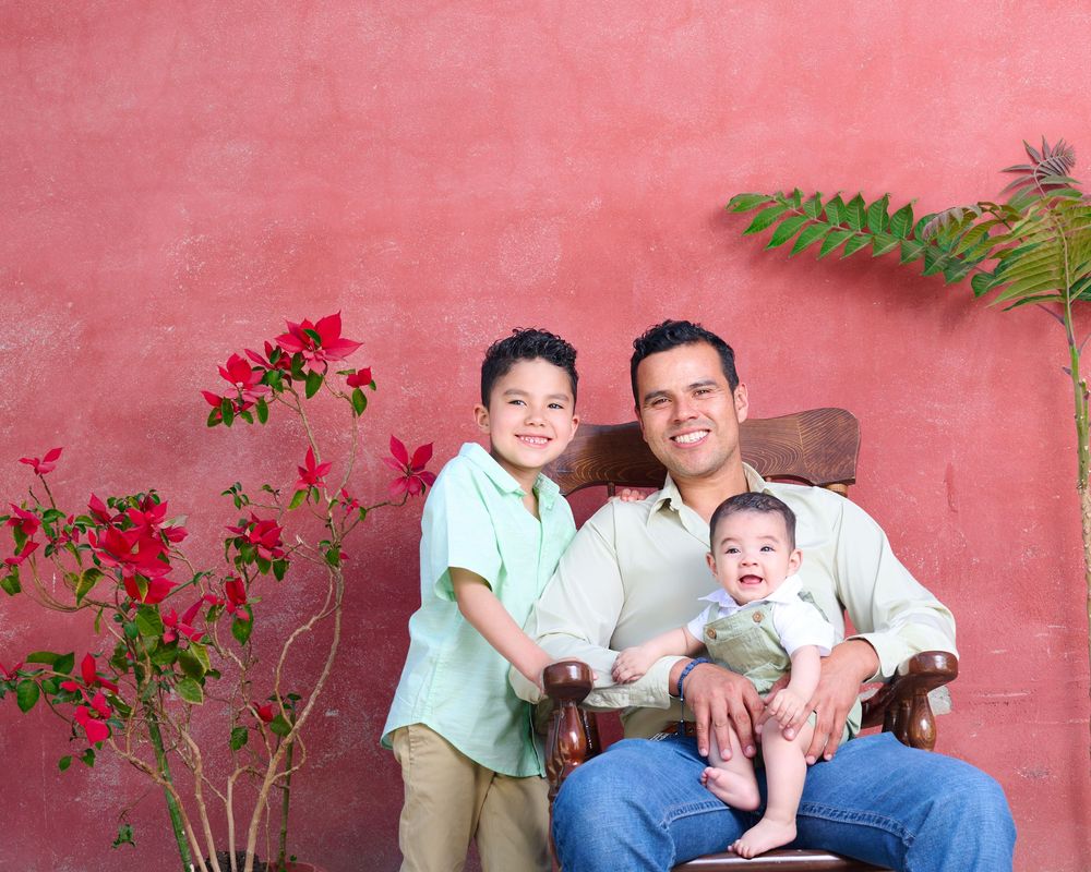 Family of three sitting together against a pink wall with red bougainvillea flowers and fern leaves.