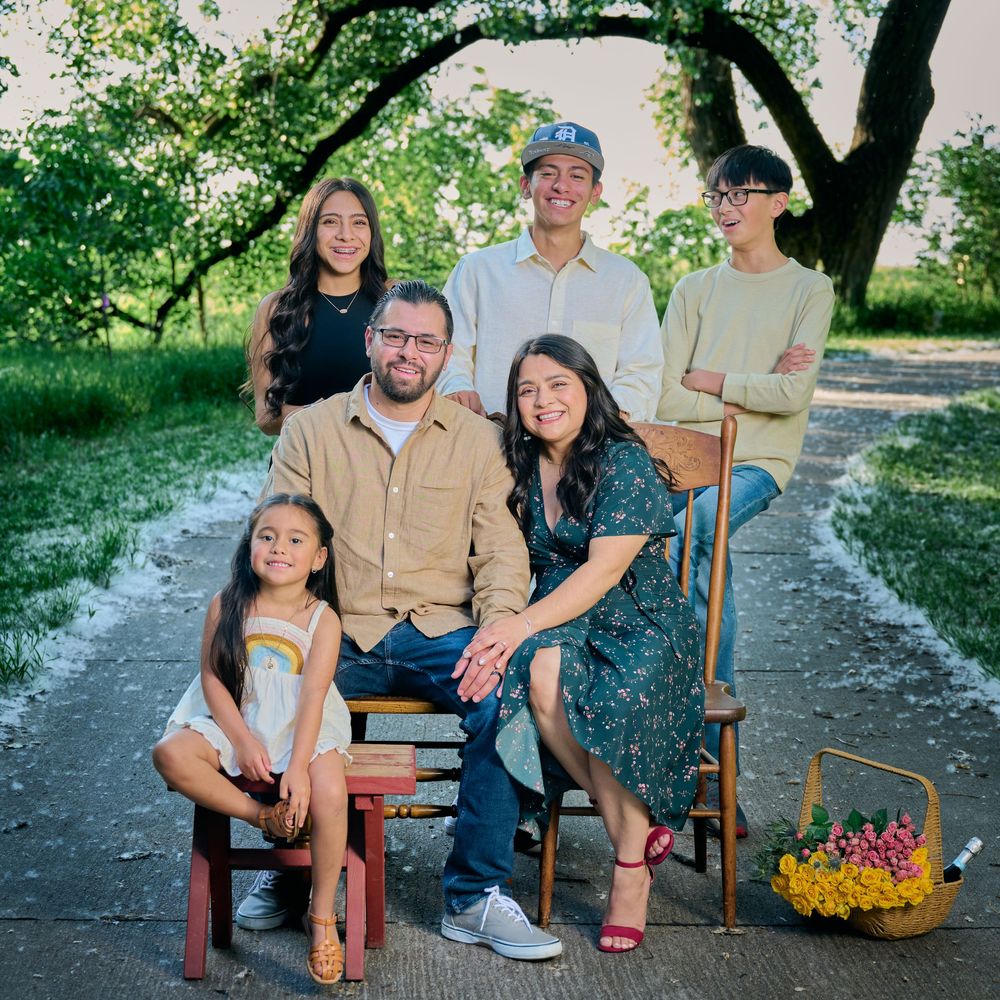 Family enjoys casual outdoor photoshoot while sitting on rustic wooden chairs surrounded by greenery and flowers.