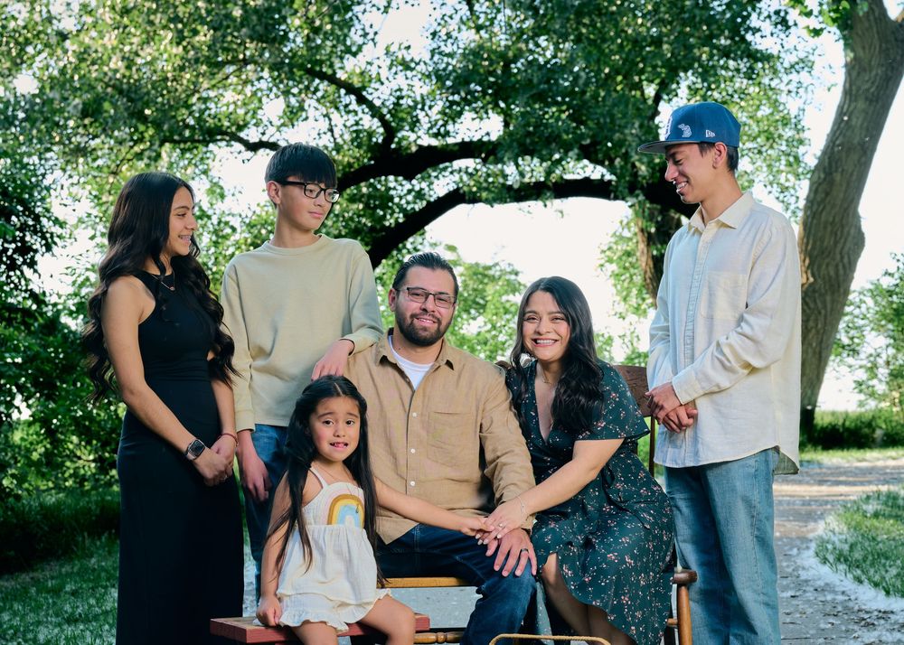 A group of family members smiling together outdoors under green trees on a sunny day.
