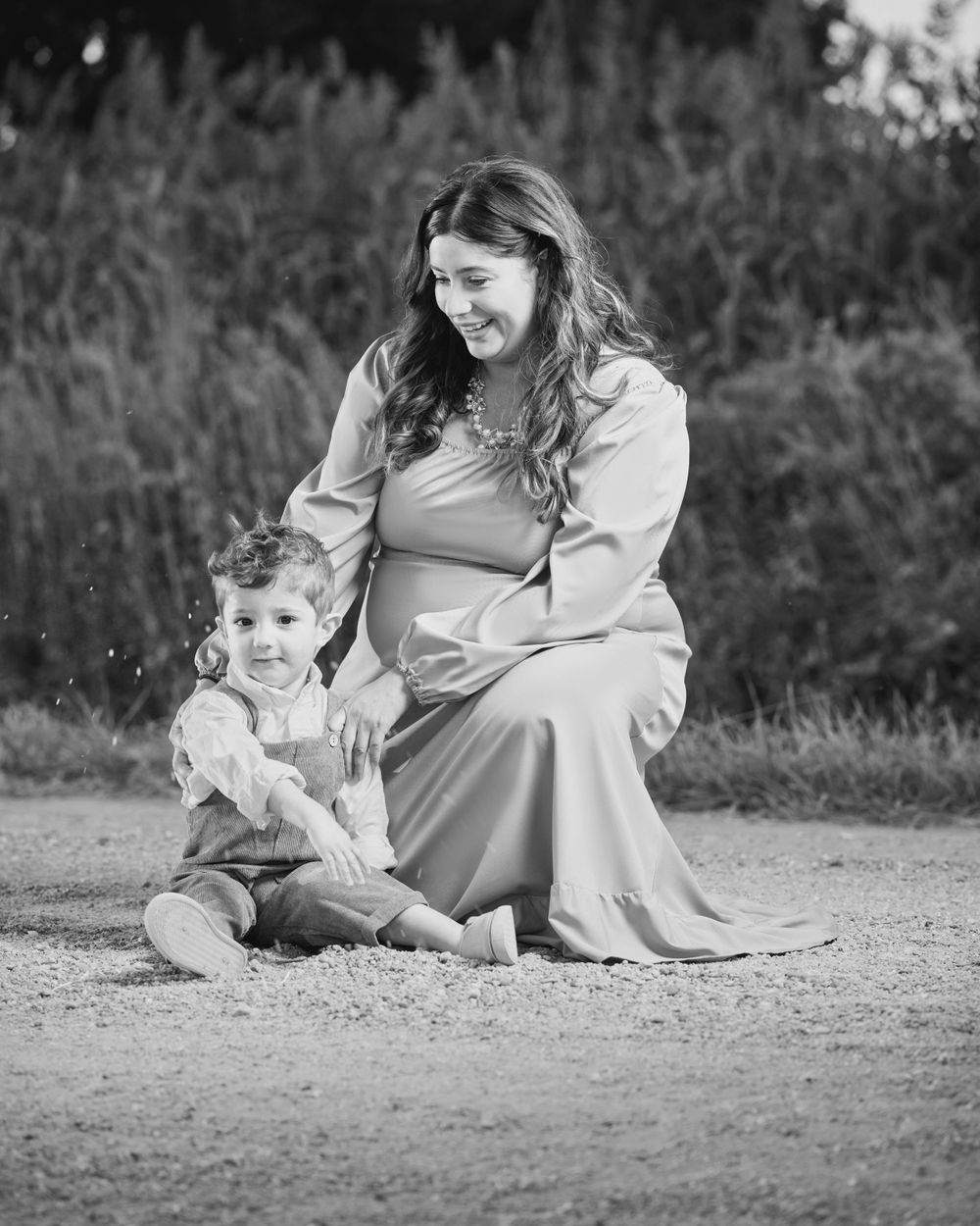 A black and white maternity portrait captured on a dirt path surrounded by tall grass.