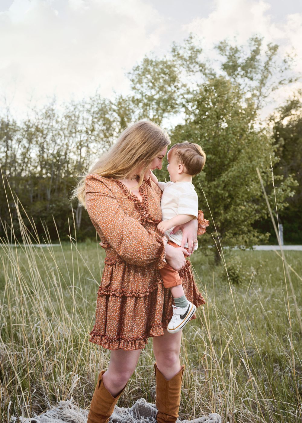 A tender outdoor moment in a field with tall grass and trees, wearing a brown ruffle dress and cognac boots.