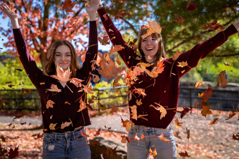 Two best friends laugh joyfully while tossing autumn leaves into the air at Hogs Back Park in Ottawa, Ontario.