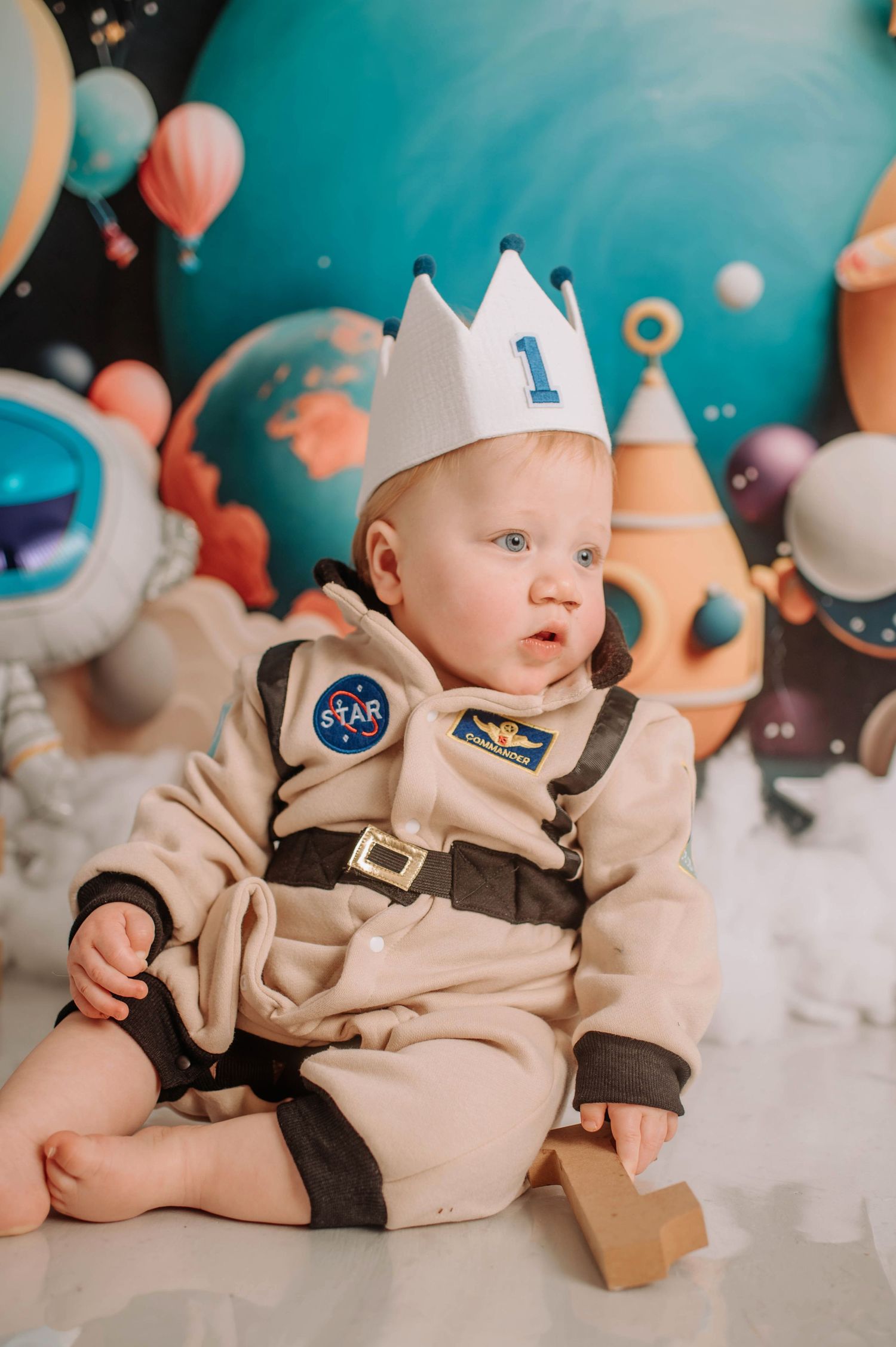 Baby wearing NASA astronaut costume and white paper crown in space-themed first birthday photoshoot setting.