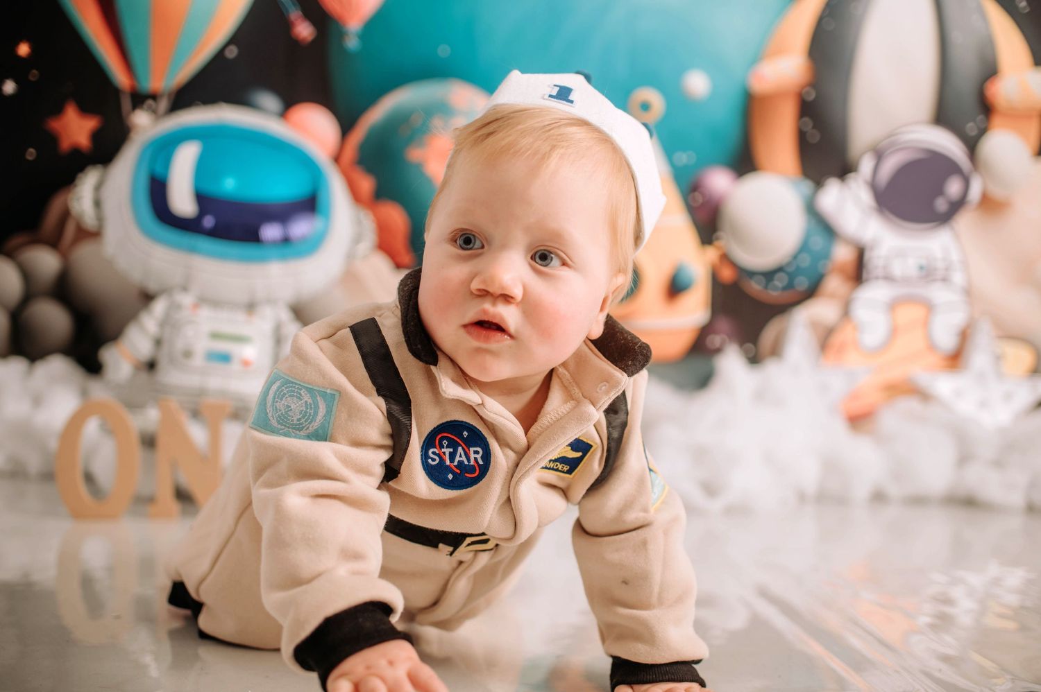 Baby in beige astronaut outfit crawls on white floor with space-themed backdrop and balloons.