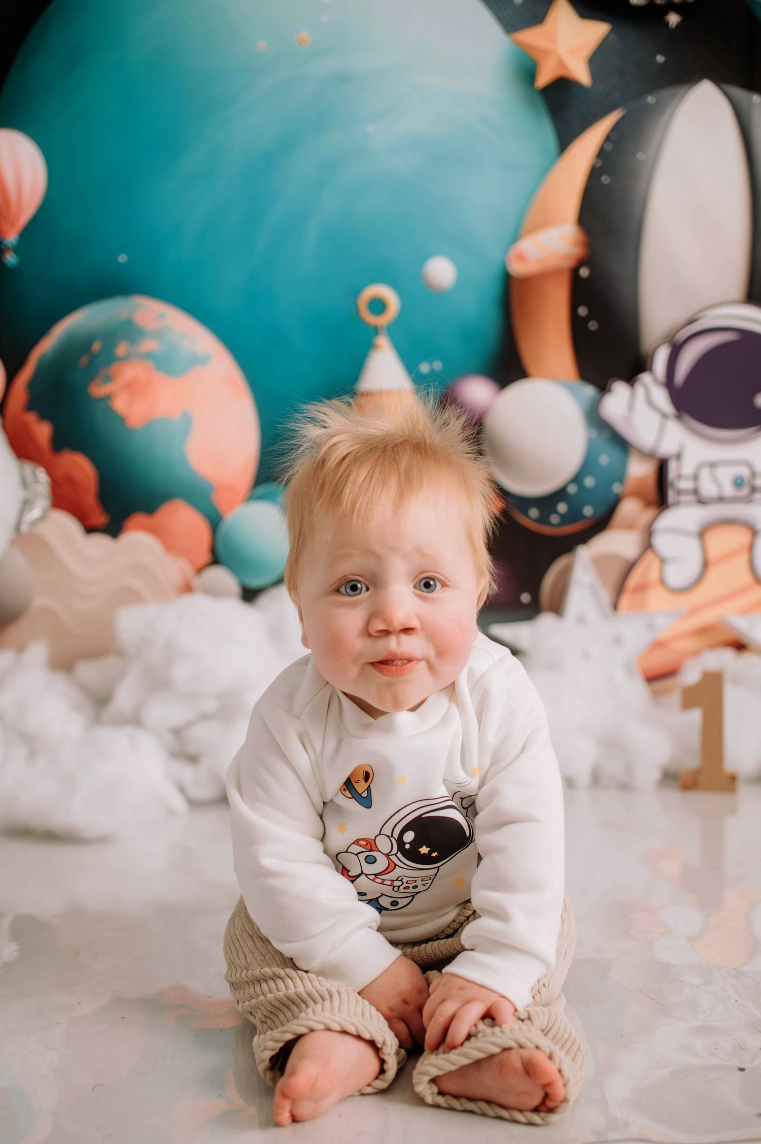 Baby in white sweater sitting on floor with space and planet themed backdrop for first birthday shoot.