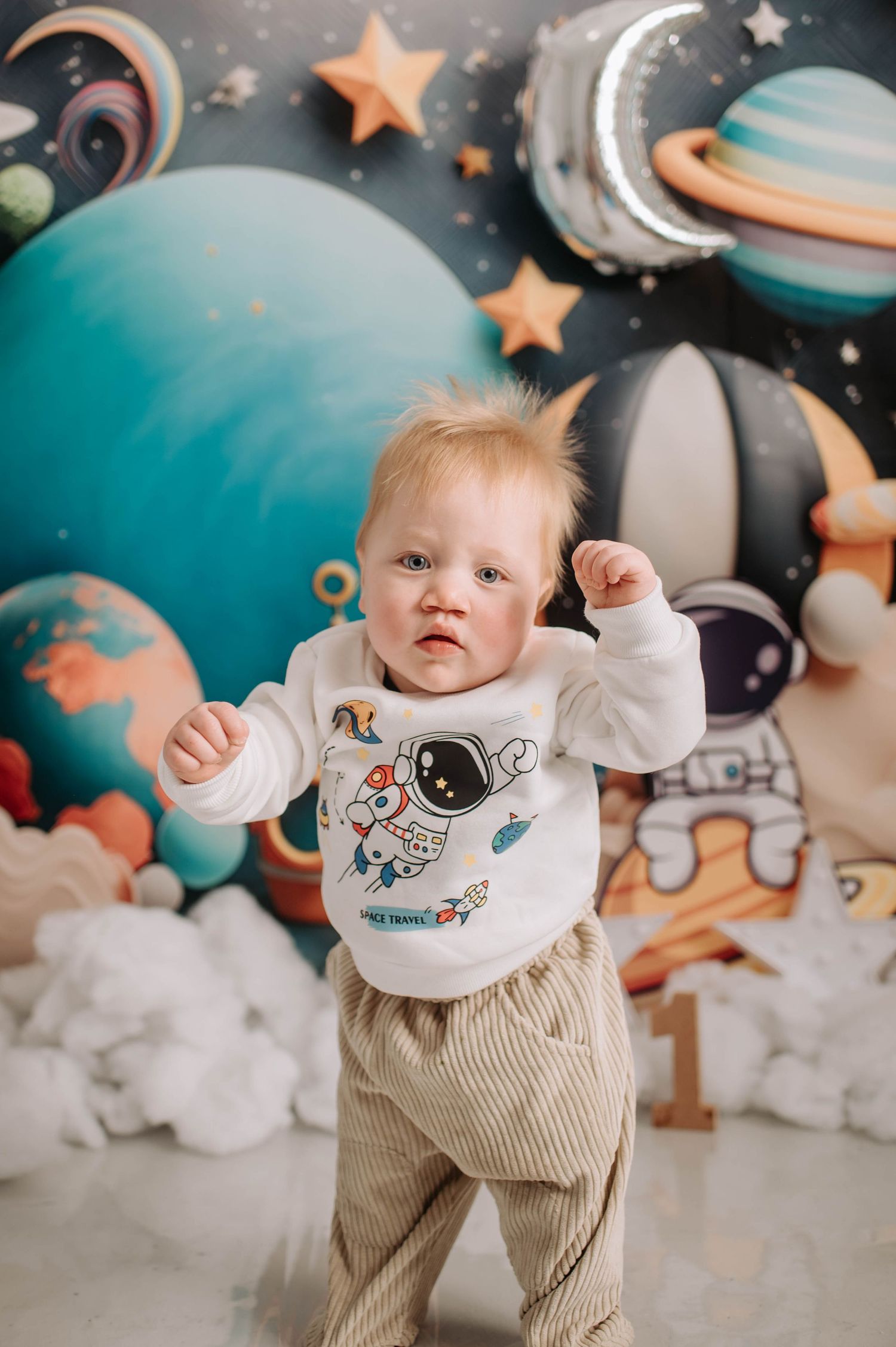 Baby standing in space-themed photoshoot wearing astronaut print sweater and beige pants.