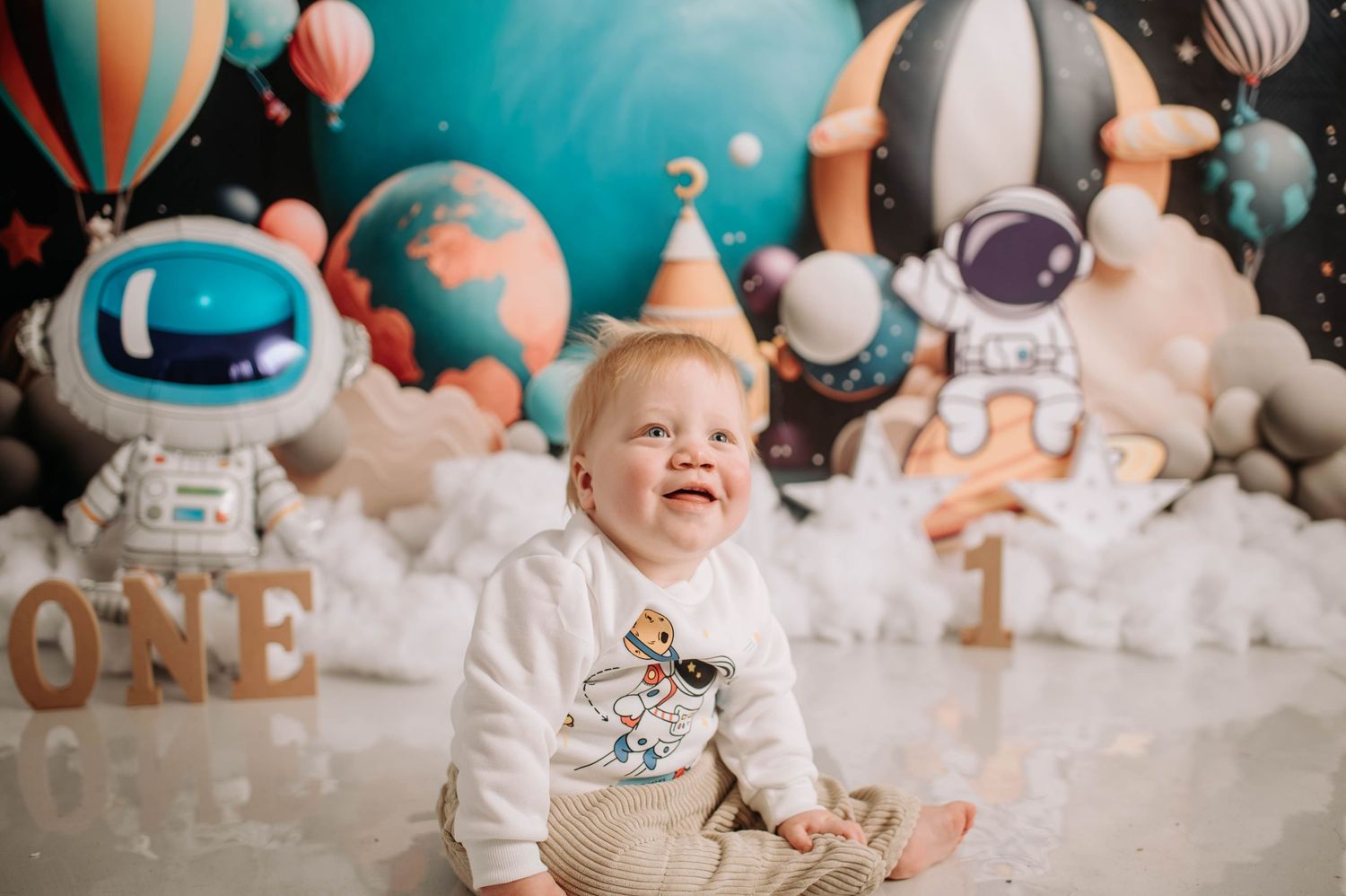 Laughing baby in party hat against cosmic backdrop with astronaut decorations.