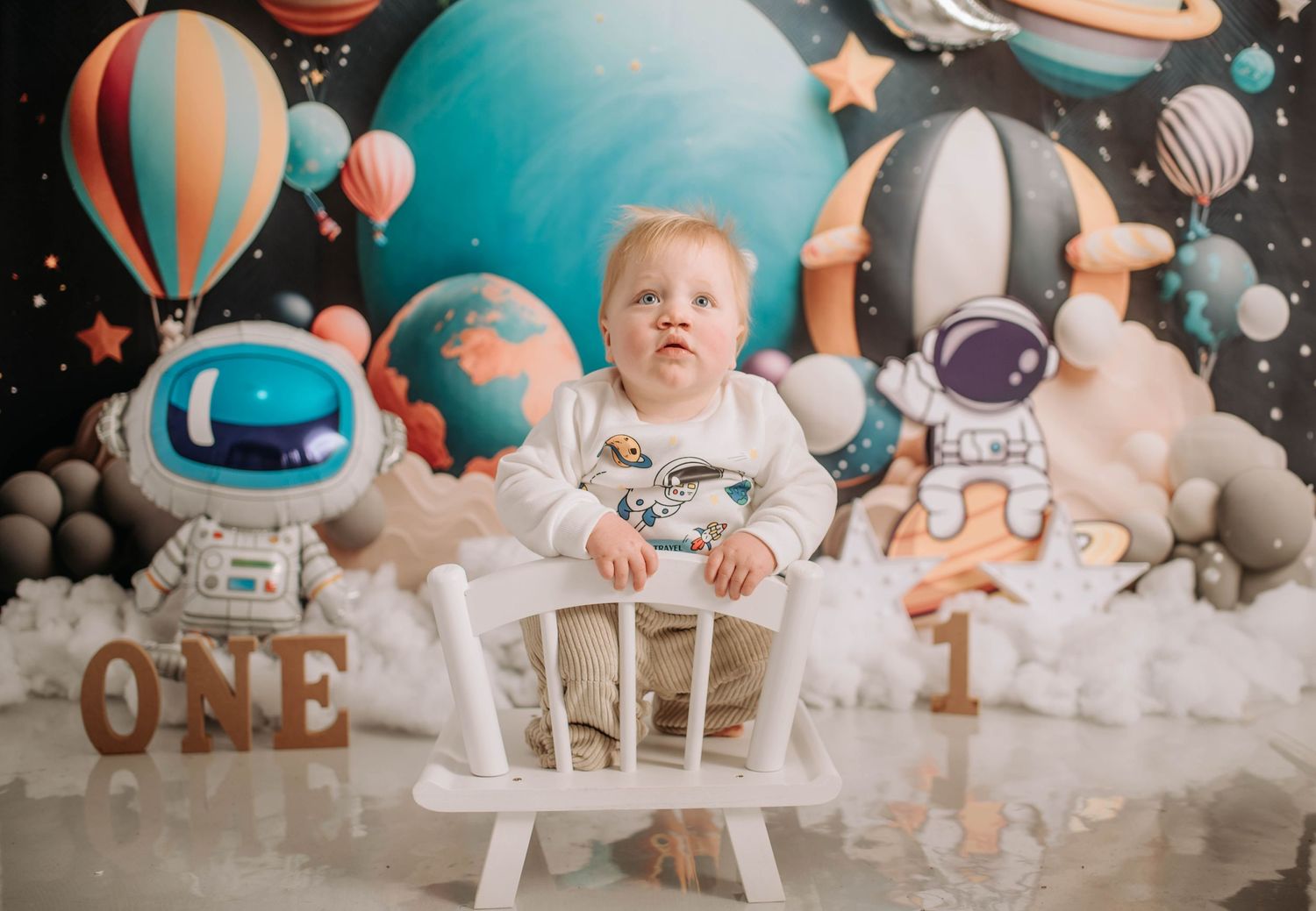 Baby on white chair surrounded by astronaut decorations and space-themed birthday setup.