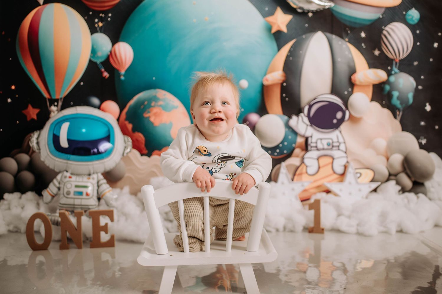 Happy baby sitting in white chair with space-themed first birthday decorations and balloons.