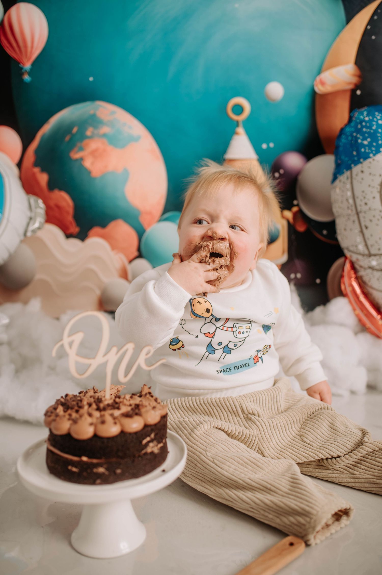 Baby with chocolate cake during space-themed first birthday photoshoot with planetary backdrop.