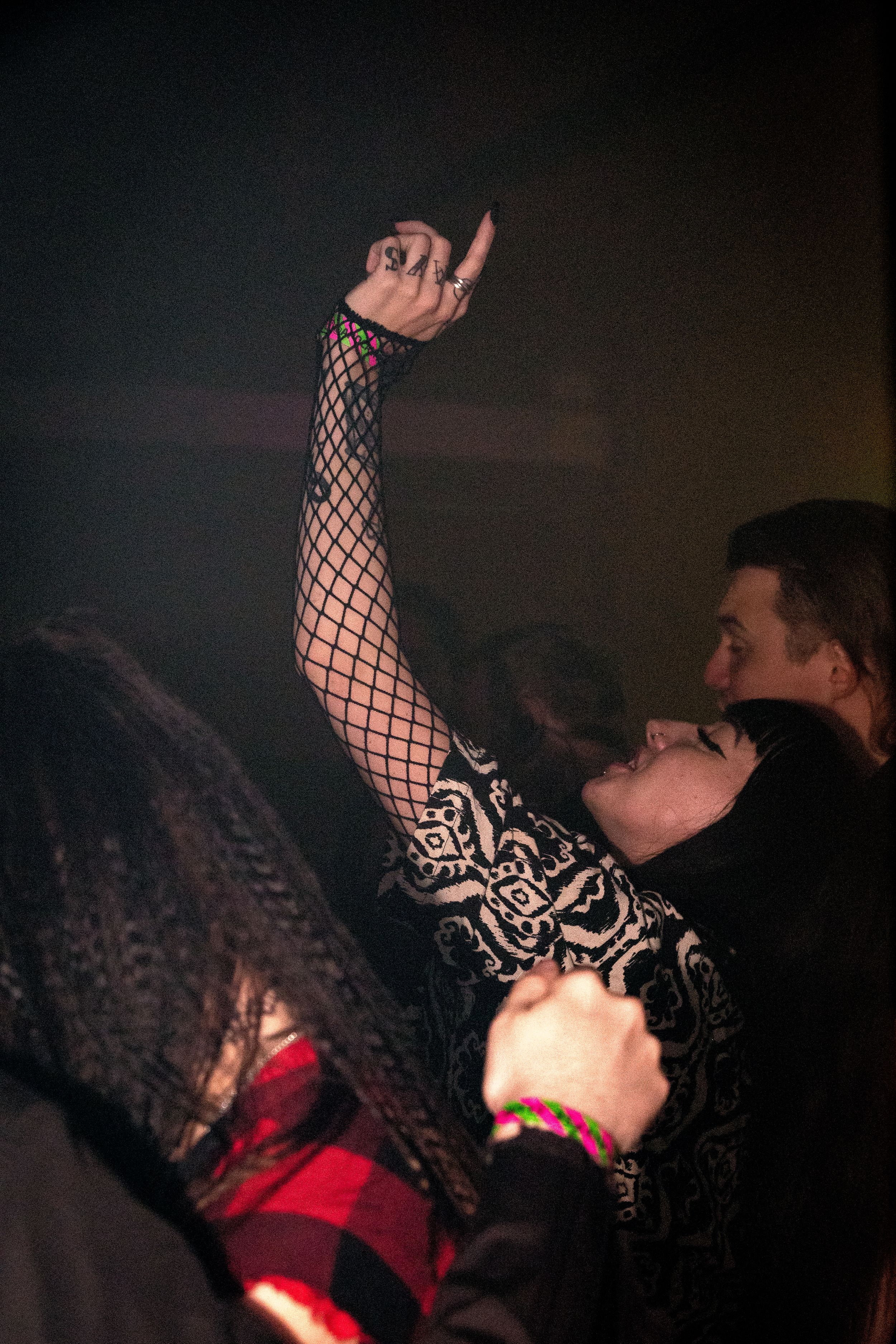 Person in fishnet sleeves dancing with arm raised at dark nightclub.