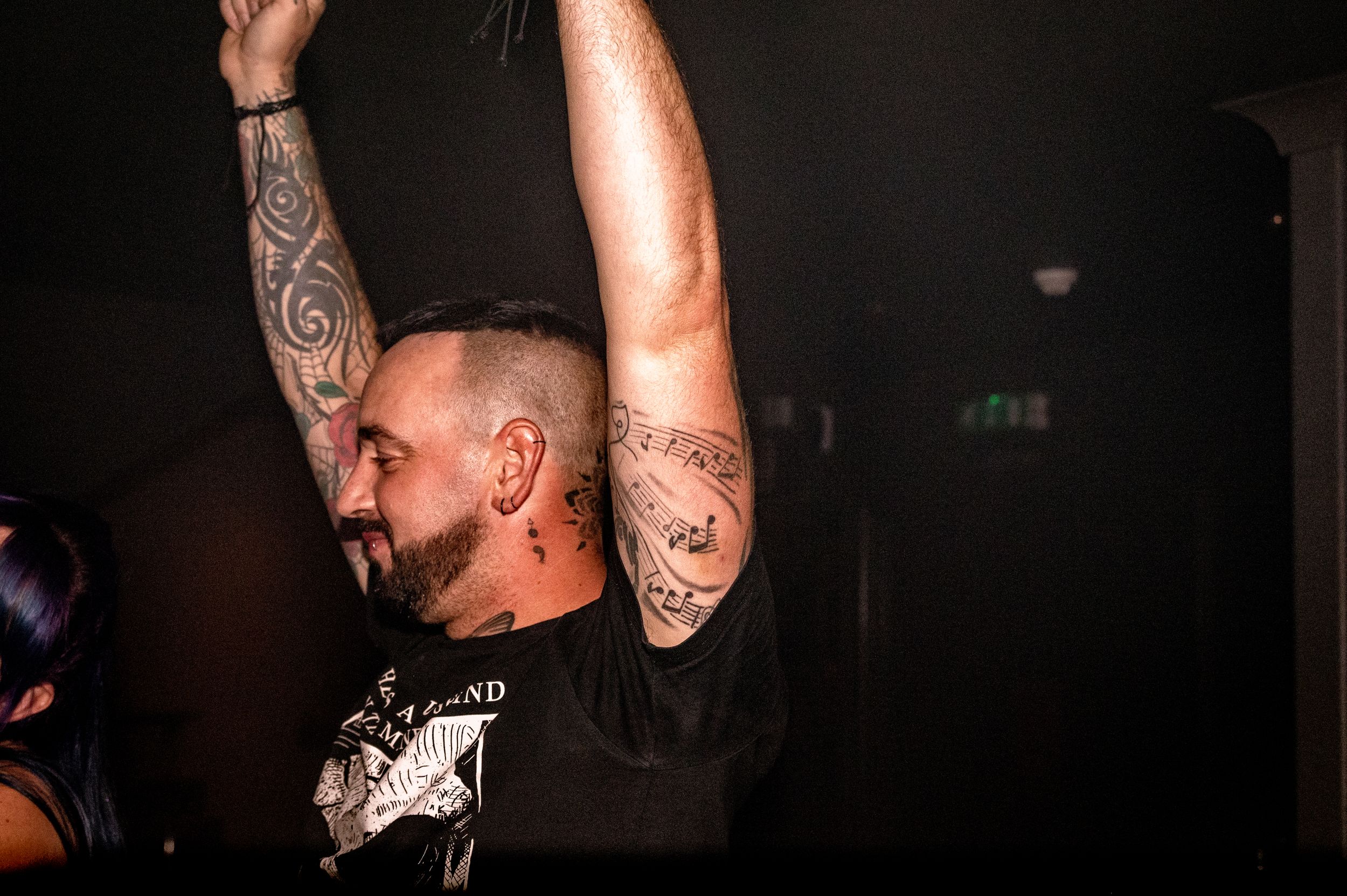 Tattooed person with raised arms dancing energetically in dark club setting.