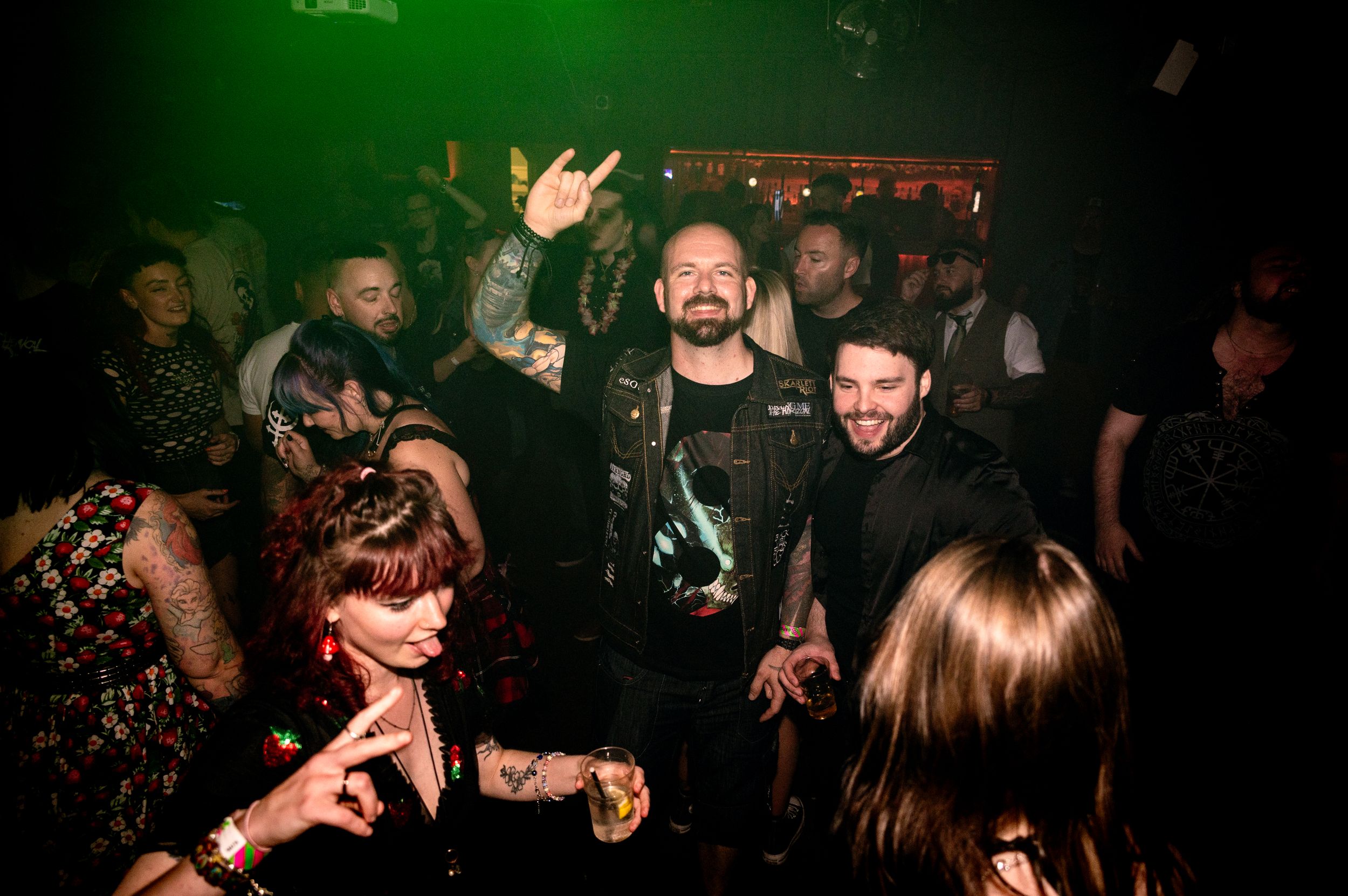 Crowd of enthusiastic concert-goers dancing and gesturing in a dimly lit nightclub with green lighting.