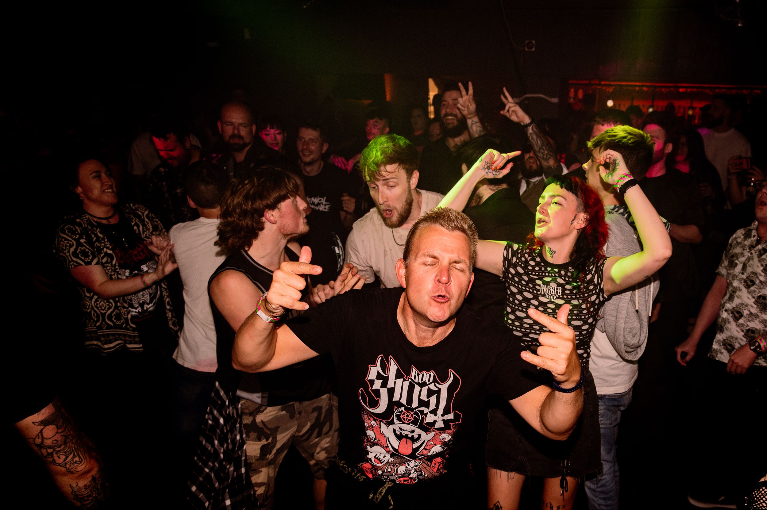 Energetic crowd at a metal concert with neon lighting and band merchandise visible.