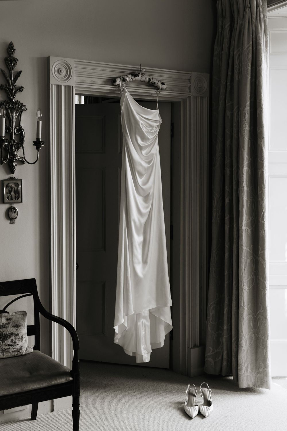 An elegant white wedding dress hanging in a doorway with shoes placed beneath.