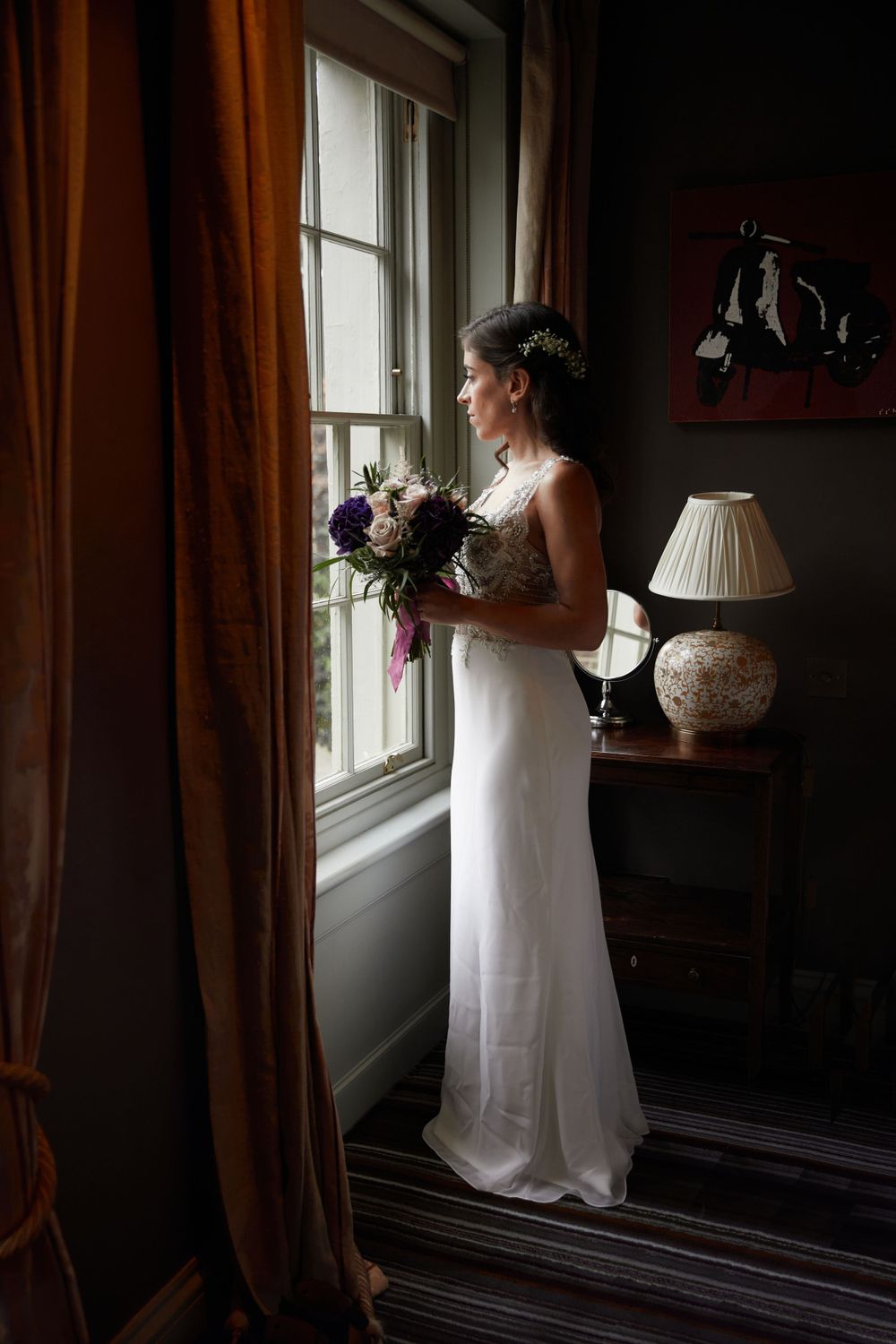Silhouette of bride holding bouquet while standing by window in elegant room.