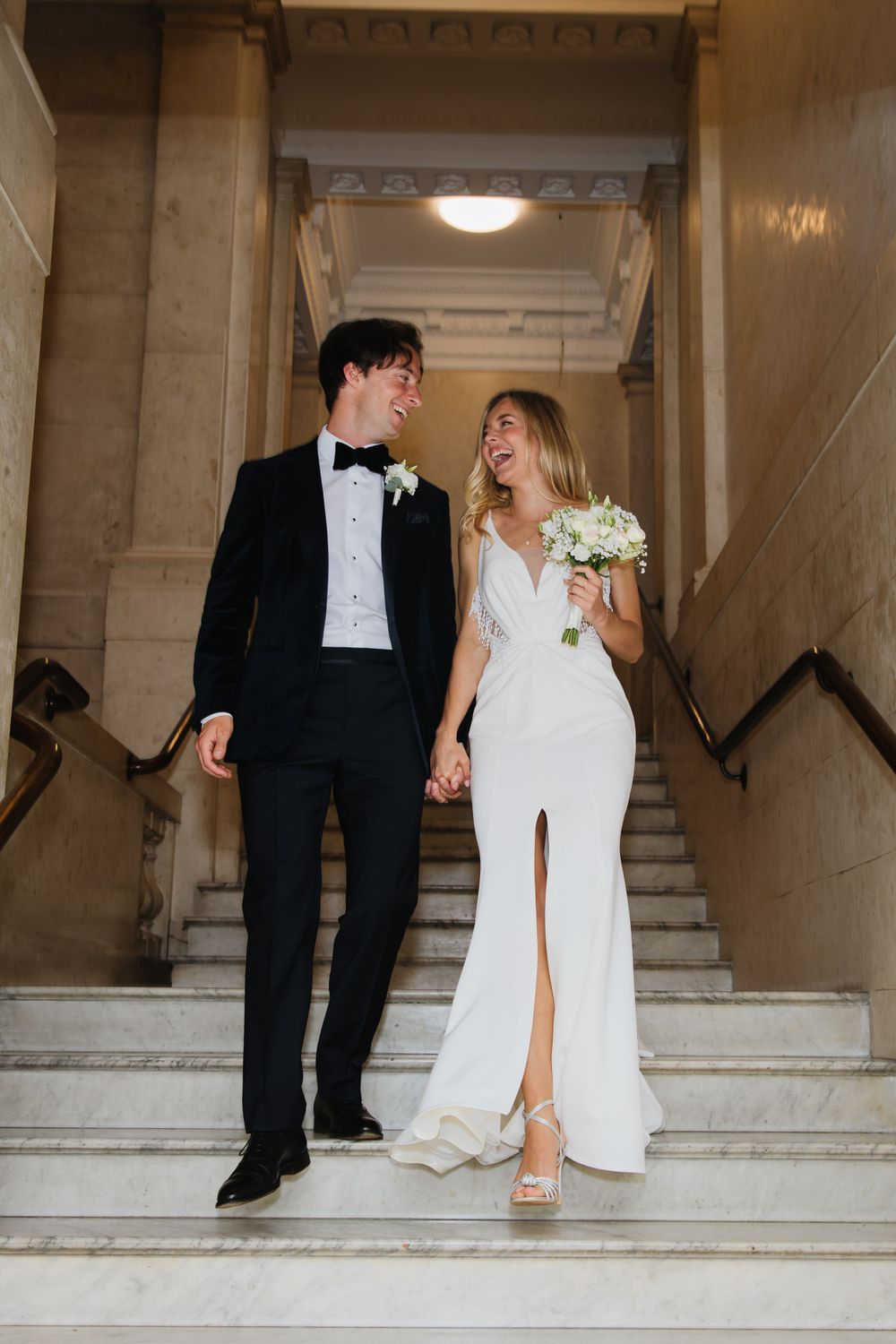 A happy couple in wedding attire standing on marble stairs in a classic building.