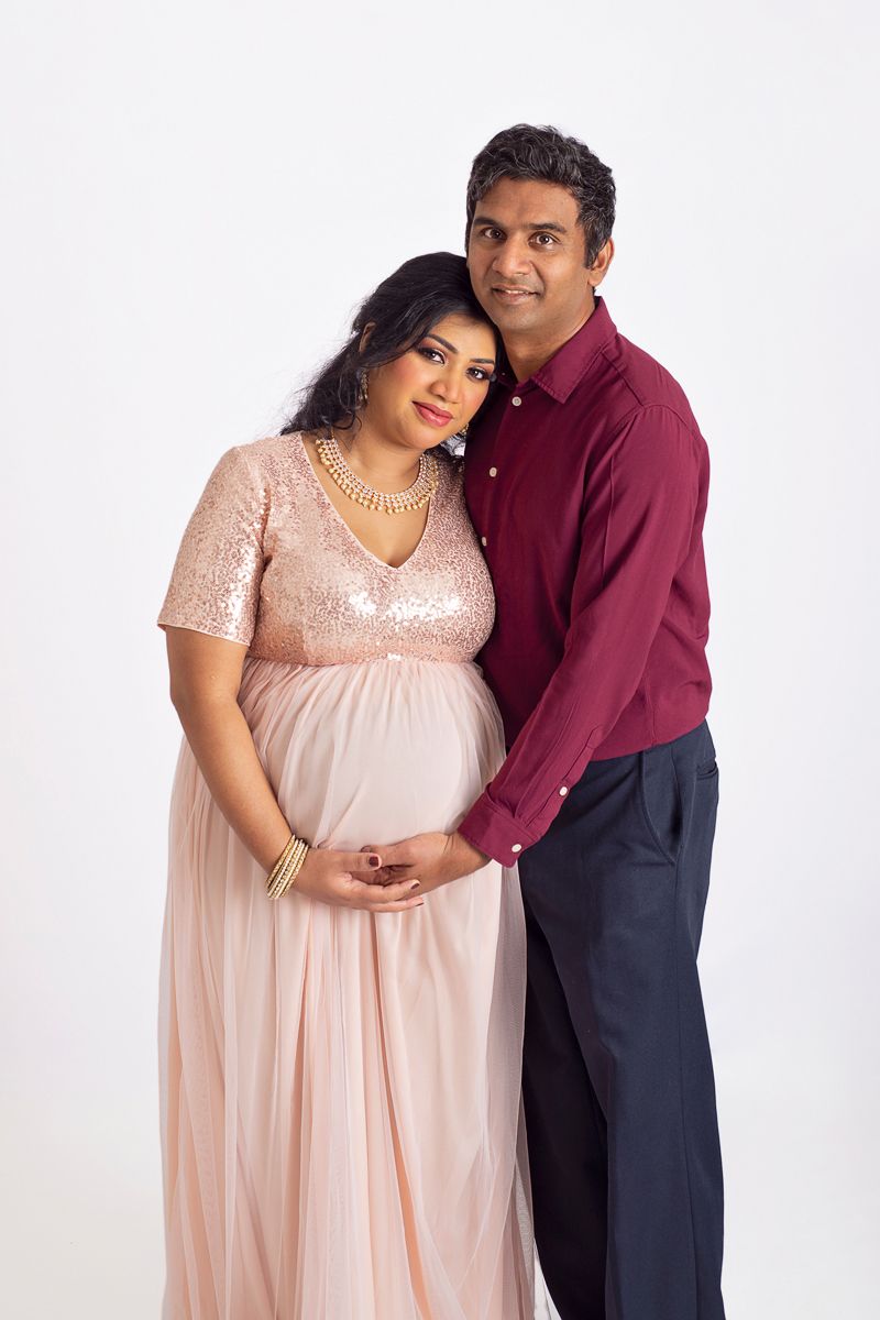 Pregnant mother in pink elegant dress is embraced by husband looking at camera