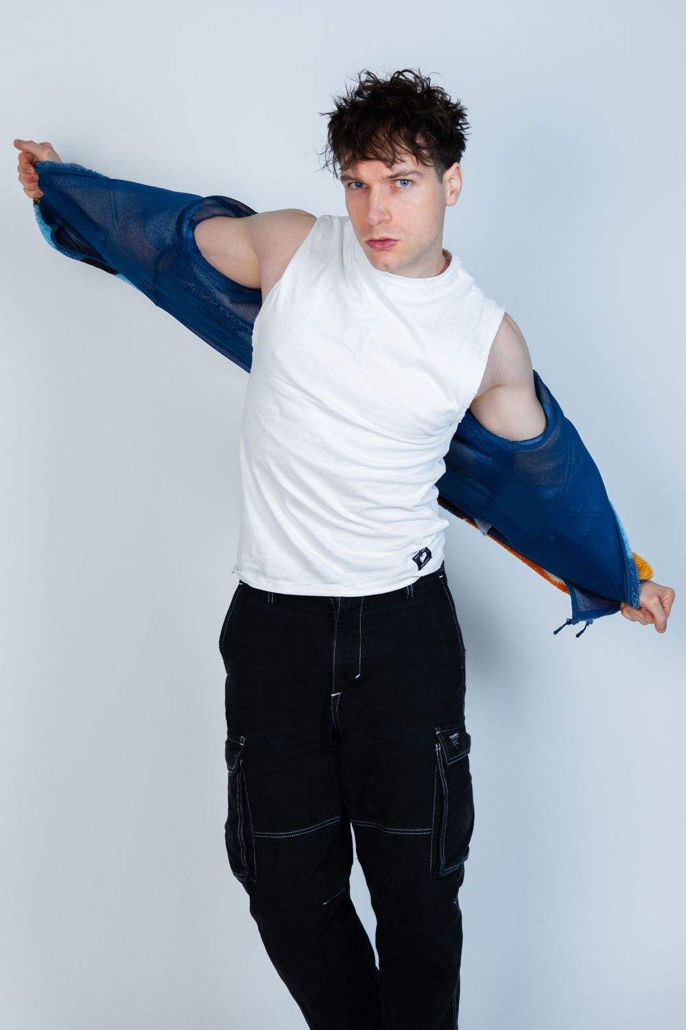 Modèle en débardeur blanc retirant une veste bleue avec un pantalon cargo noir sur fond clair.