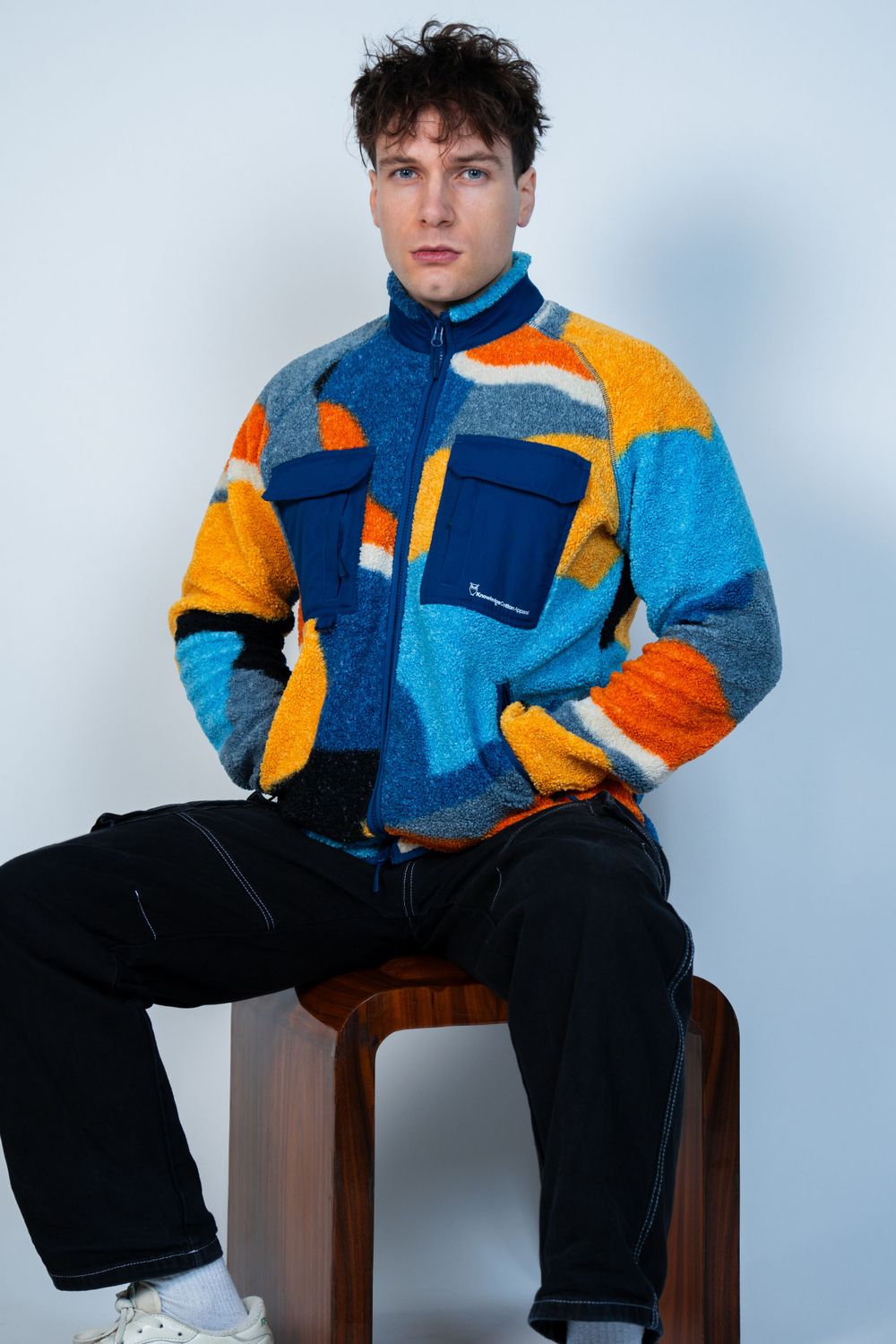 Modèle portant une veste polaire patchwork colorée dans les tons bleu et orange avec un pantalon noir sur un tabouret en bois.