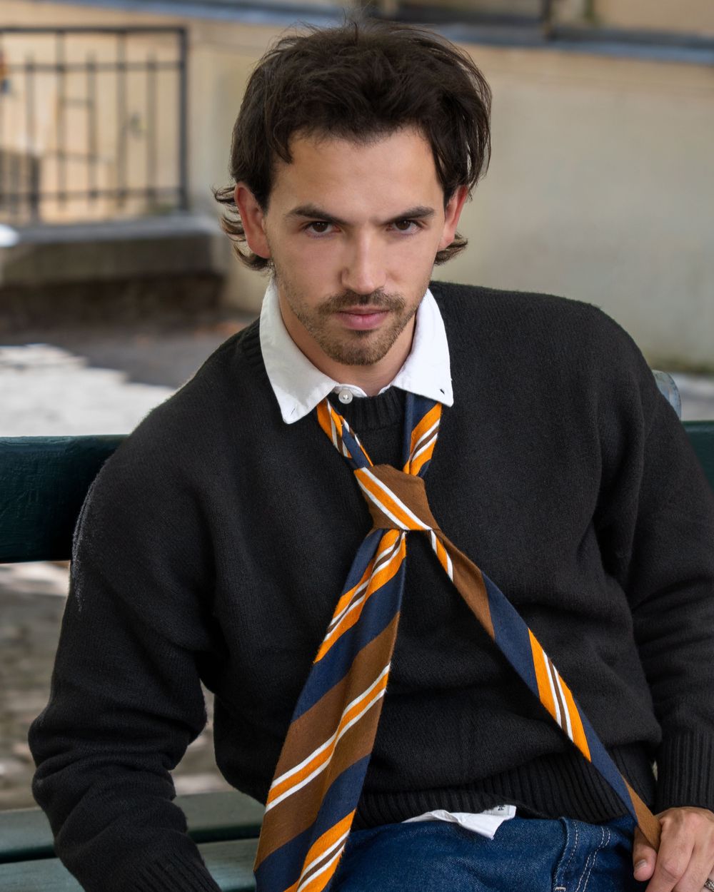 A person wearing a dark sweater and striped school tie leans casually against a wall.