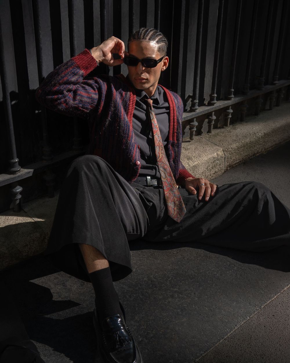 Stylish man in dark trousers, red cardigan, tie, and sunglasses sitting outdoors on steps in moody lighting.