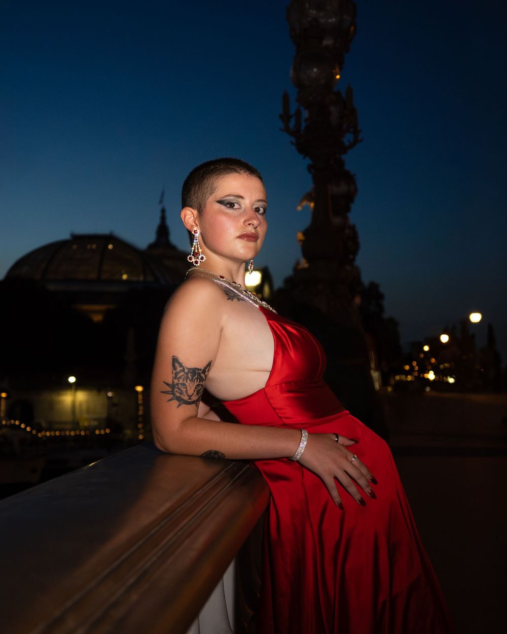 Woman in red gown with shaved head and tattoos poses on bridge at night.