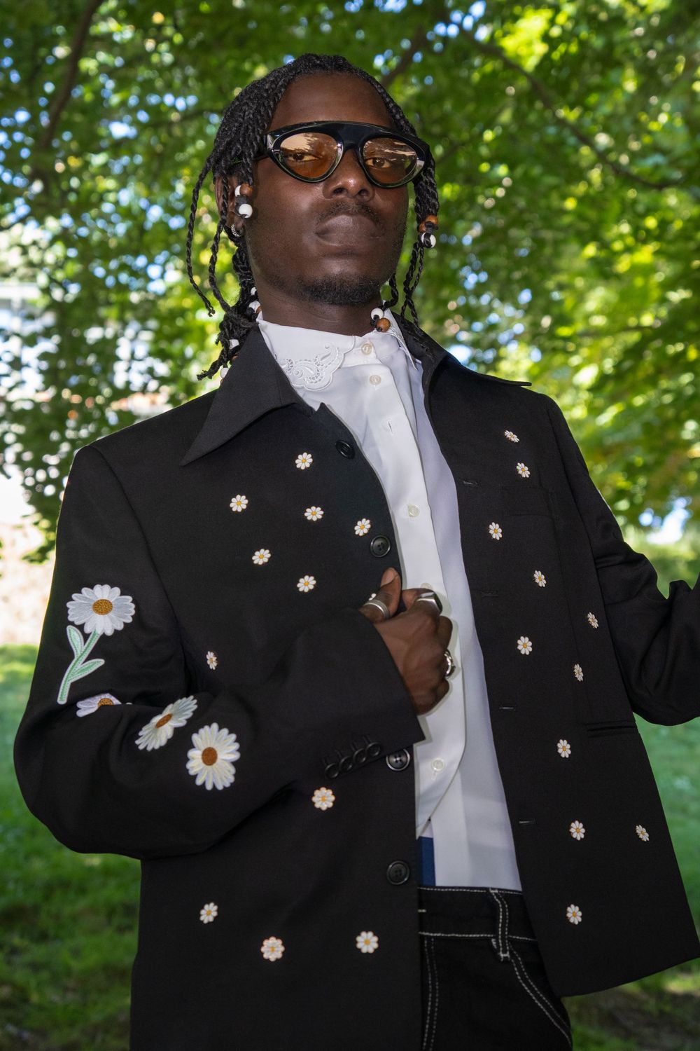 A person in a black jacket with daisy flower embellishments and white shirt poses playfully in an outdoor setting.
