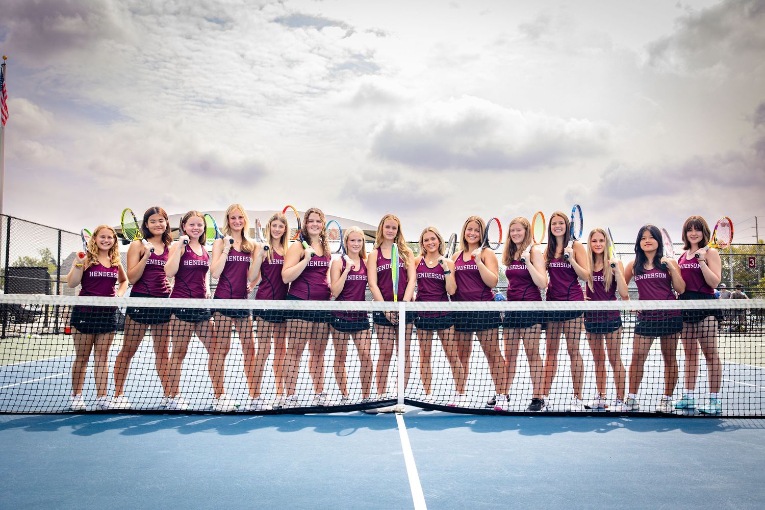 Team photo of athletes lined up on blue tennis court under cloudy sky.