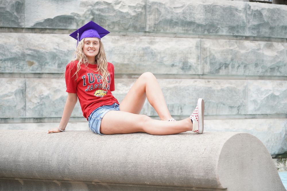 Graduate in red shirt and purple cap lounges on concrete ledge.