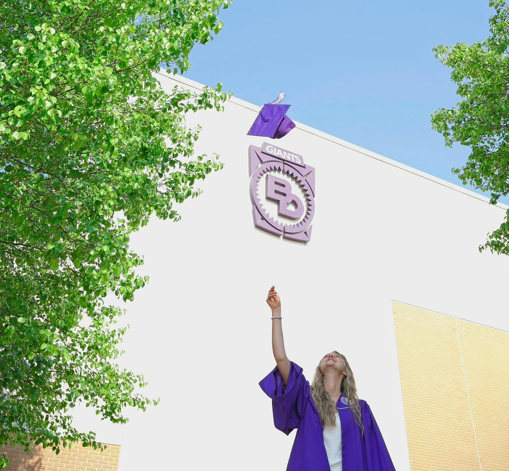 Graduate in purple gown tosses cap in front of white building with school emblem.