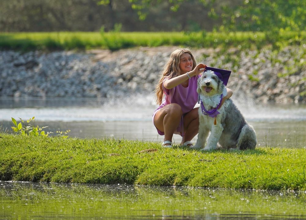 Person in casual attire crouches next to a fluffy white and gray dog by a pond.