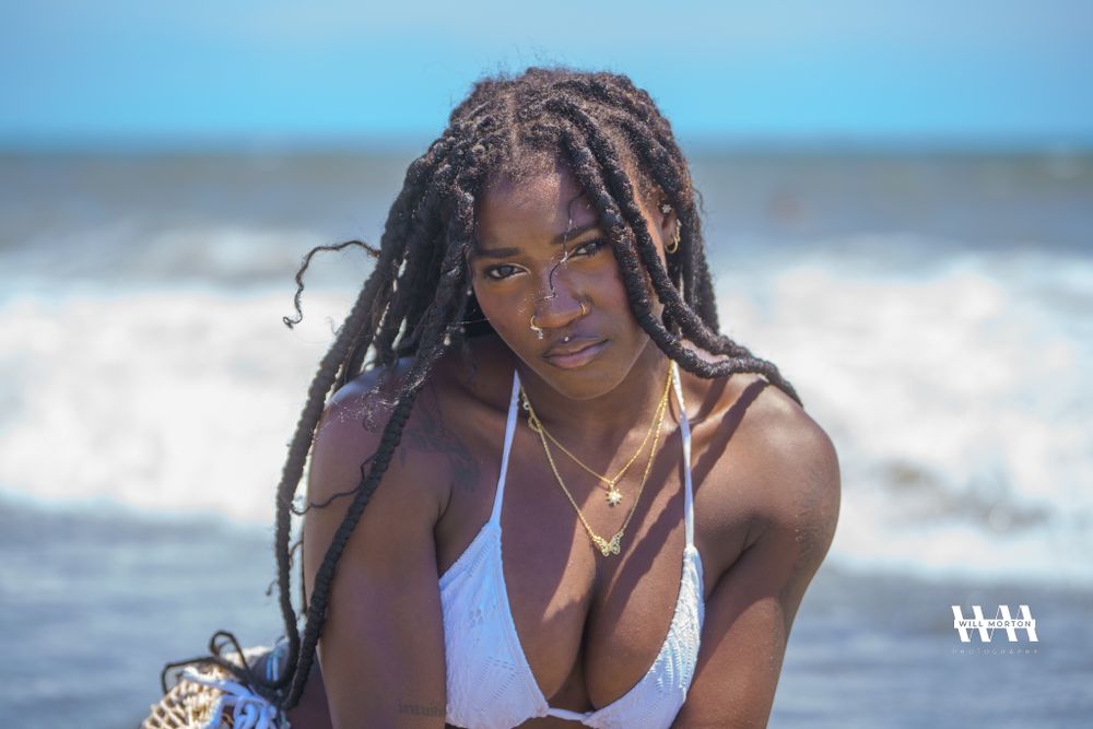 Model in a white swimsuit poses confidently against ocean waves and blue sky backdrop at the beach.