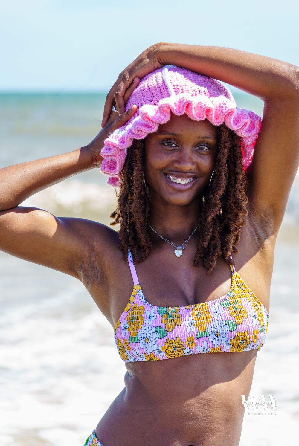 Summer beach portrait with pink ruffle hat and colorful patterned bikini against ocean backdrop.