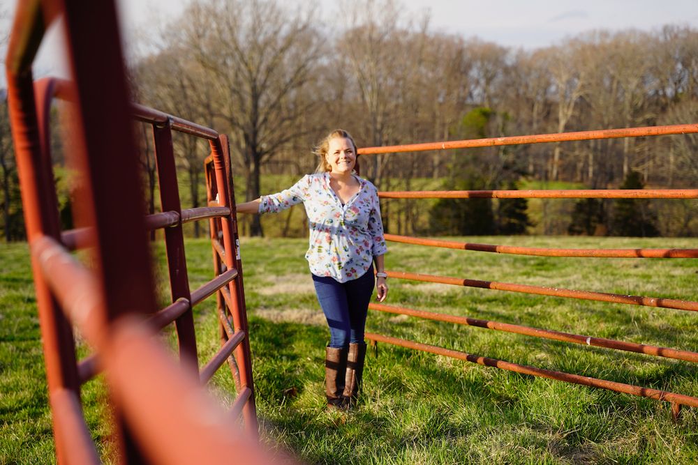 Person posing playfully by red metal fence gates in a pastoral country setting.