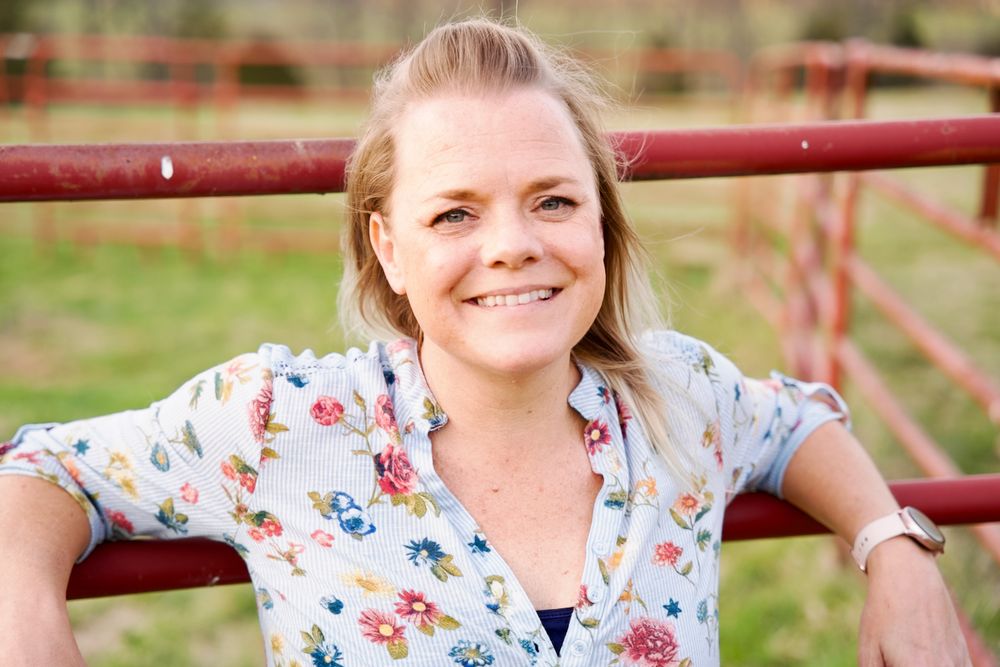Person in floral print top smiles warmly while posing near red farm fence.