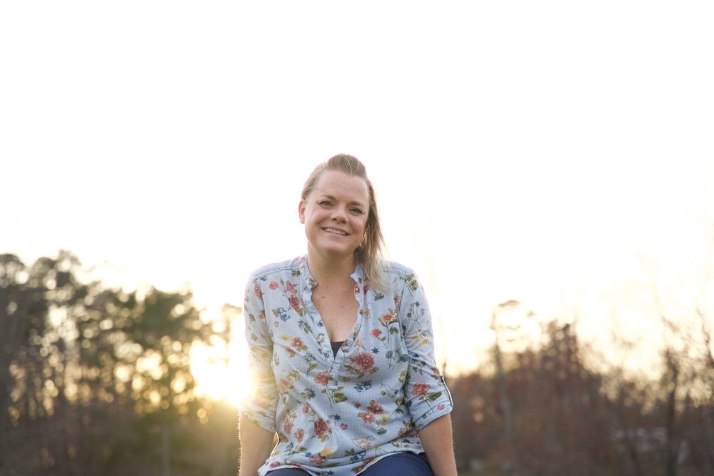 Person in floral dress poses against scenic sunset backdrop in rural setting.
